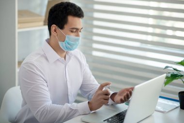 Beyaz gömlekli bir adam laptopunu dezenfekte ediyor ve ciddi görünüyor.