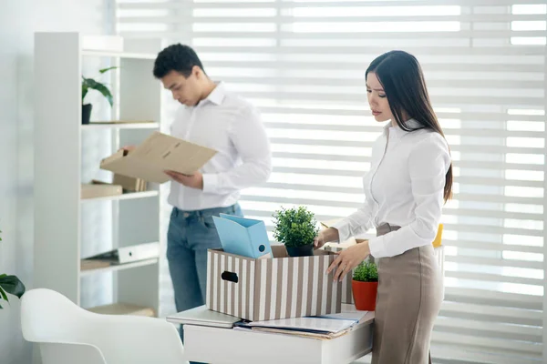 Jong brunette vrouw staan aan haar bureau, het verzamelen van haar persoonlijke spullen, haar mannelijke collega staan achter — Stockfoto