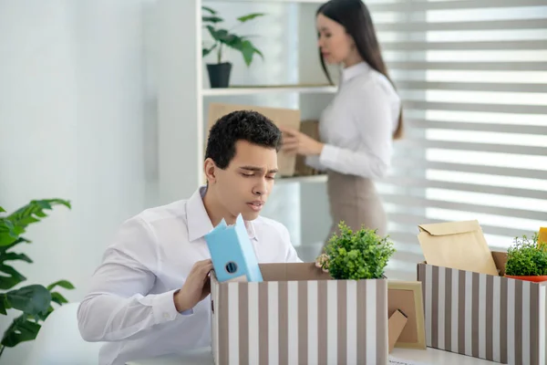 Jonge brunette man zitten aan zijn bureau, het zetten van zijn persoonlijke spullen in doos, zijn vrouwelijke collega staan achter — Stockfoto