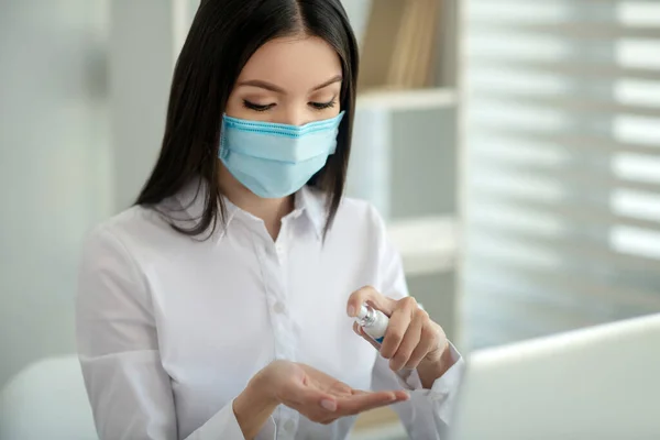 Mujer joven en una máscara facial rociando desinfectante en sus manos — Foto de Stock