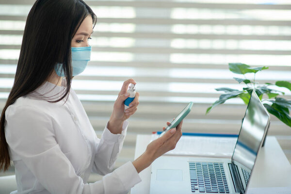 Молодая женщина в защитной маске дезинфицирует свой смартфон
