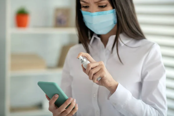 Mujer joven en una máscara facial rociando desinfectante en su teléfono inteligente — Foto de Stock