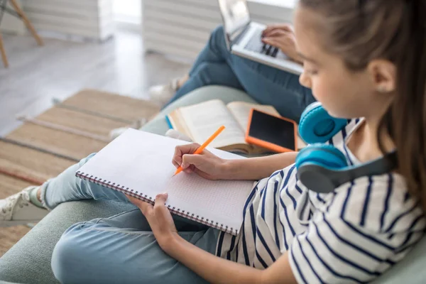 Mädchen mit Kopfhörern schreibt in Notizbuch, während sie auf dem Sofa sitzt. — Stockfoto