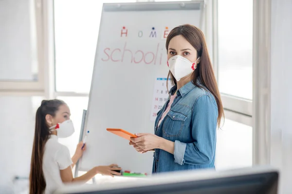 Frau mit Schutzmaske und Tablet in der Hand, Mädchen an der Tafel. — Stockfoto