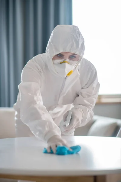 Жінка в білому робочому одязі і захисних рукавичках виглядає концентрованою під час дезінфекції поверхні столу — стокове фото