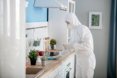 Beyaz iş elbiseli ve koruyucu gözlüklü kadın mutfağı dezenfekte ediyor.