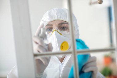 Beyaz elbiseli ve solunum cihazlı kadın pencereyi dezenfekte ediyor.