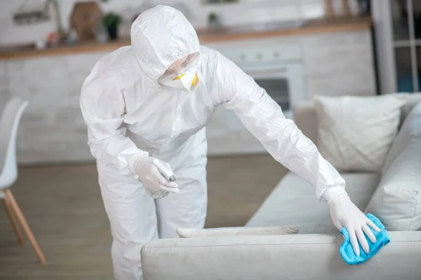 Mujer en ropa de trabajo blanca y guantes protectores desinfectando muebles —  Fotos de Stock