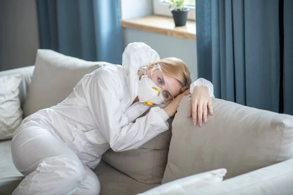 Mujer rubia en ropa de trabajo blanca y respirador descansando en el sofá — Foto de Stock
