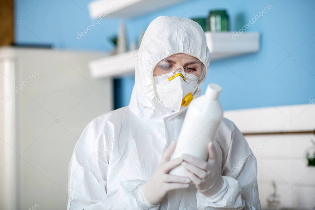 Woman in white workwear and gloves holding a bottle with antiseptic liquid