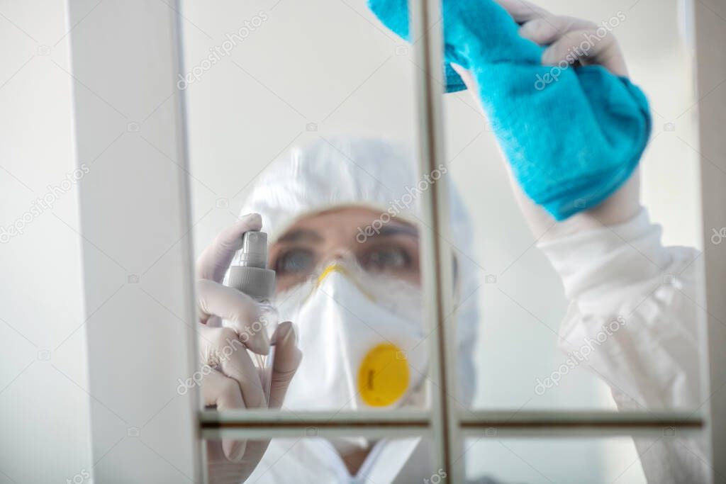 Woman in white workwear and respirator spraying disinfecting liquid