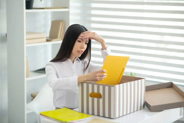 Jong brunette vrouw zitten aan haar bureau met doos van persoonlijke spullen en aanraken van haar voorhoofd — Stockfoto