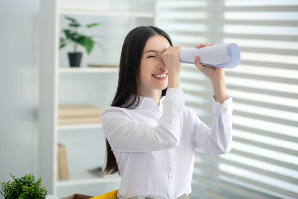 Молодая брюнетка женщина смотрит через бумажную трубку, улыбаясь
