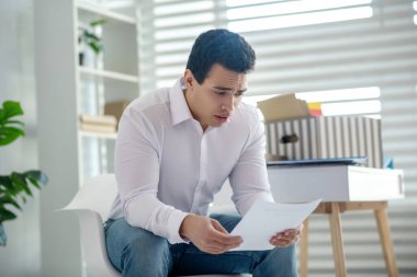 Genç, esmer bir erkek masasında oturuyor, elinde kağıt parçasıyla.