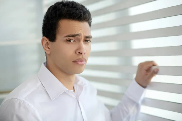 Upset young brunette male standing at the window, touching blinds, looking sideways — Stock Photo, Image