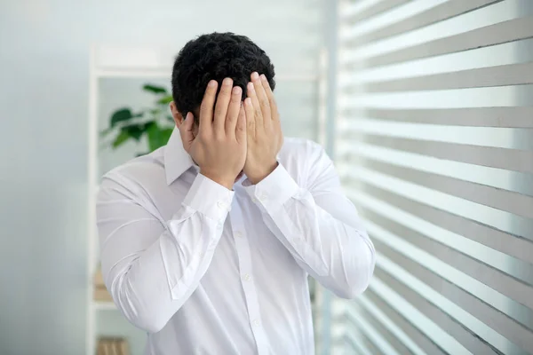 Verstoorde jonge brunette man staan aan het raam, het bedekken van zijn gezicht met zijn handen — Stockfoto