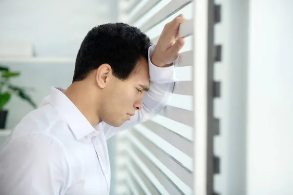 Upset young brunette male standing at the window, leaning his arm and head on blinds — Stock Photo, Image