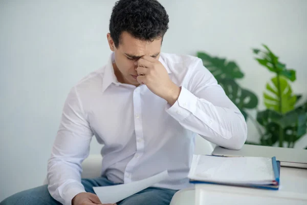 Upset young brunette male sitting at his desk, holding piece of paper, touching his nose bridge — Stock Photo, Image