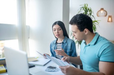 Esmer erkek ve siyah saçlı kadın masada oturuyor, evrakları kontrol ediyor.