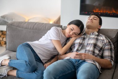 Koyu saçlı kadın başını esmer erkek omzuna yaslıyor, ikisi de kanepede dinleniyor.