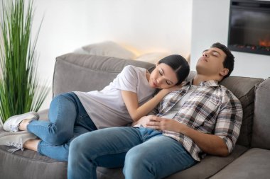 Koyu saçlı kadın başını esmer erkek omzuna yaslıyor, ikisi de kanepede dinleniyor.