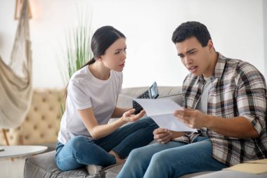 Esmer erkek, elinde kağıt, koyu saçlı kadın elinde kalküteriyle oturuyor, ikisi de stresli görünüyor.