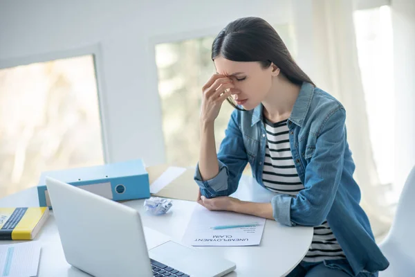 Femme aux cheveux bruns bouleversée assise à son bureau avec demande de prestations de chômage, touchant son pont du nez — Photo