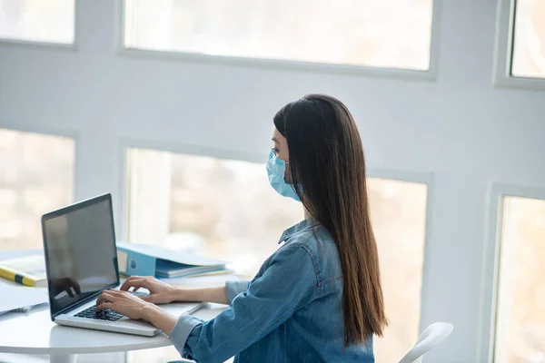Femme aux cheveux bruns assise à son bureau, tapant sur un ordinateur portable, portant un masque de protection — Photo