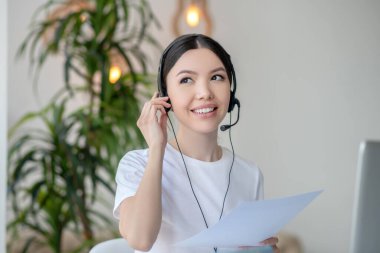 Esmer kadın kulaklık takıyor, elinde kağıtlar tutuyor, telefon alıyor.