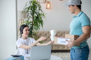 Koyu saçlı kadın, esmer erkek kuryeden yiyecek kutuları alıyor, elinde paketler var.