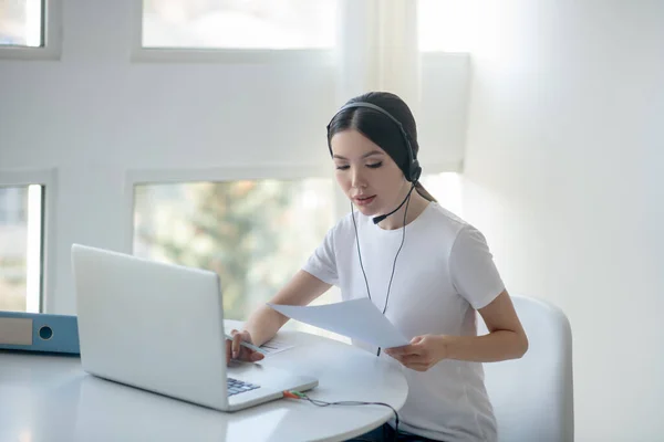 Morena mujer escribiendo en el ordenador portátil, usando auriculares, sosteniendo papeles — Foto de Stock