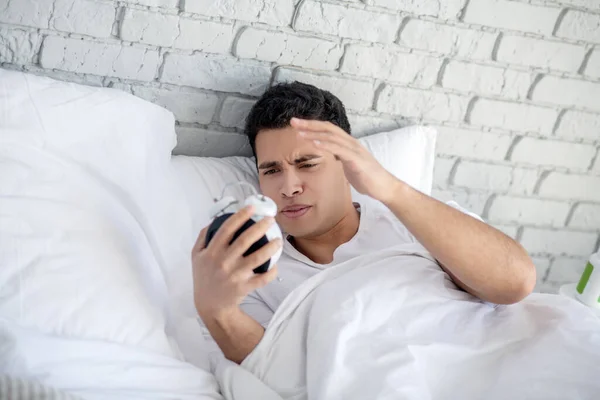 Brunette laki-laki berbaring di tempat tidur, memegang alarm-jam — Stok Foto