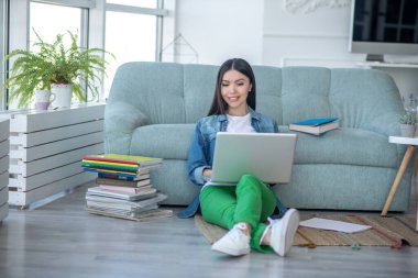 Yeşil pantolonlu genç bayan yerde oturuyor ve uzaktan çalışıyor.