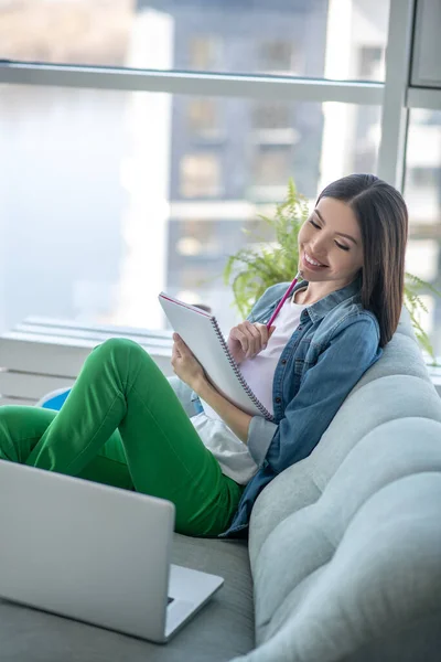 Mujer joven en una chaqueta vaquera sentada en el sofá y preparándose para los exámenes — Foto de Stock