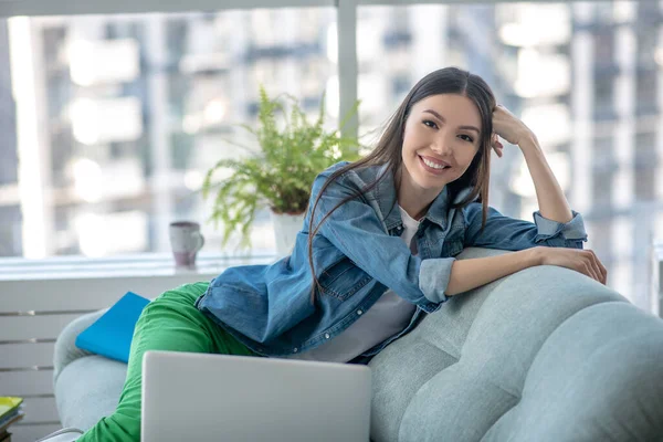 Giovane donna in giacca di jeans seduta sul divano e sorridente — Foto Stock