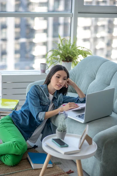 Mujer joven en pantalones verdes sentada en el suelo y escuchando un webinar — Foto de Stock