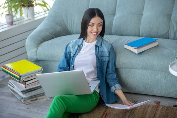 Mujer joven en pantalones verdes sentado en el suelo con un ordenador portátil y muchos libros — Foto de Stock