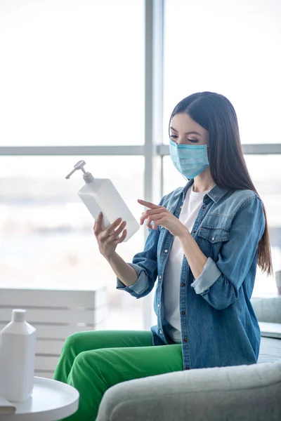 Mujer joven con una máscara protectora sosteniendo una botella con desinfectante — Foto de Stock