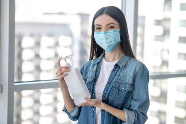 Mulher de cabelos escuros em uma máscara facial segurando uma garrafa com desinfetante para casa — Fotografia de Stock