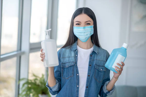 Mujer de cabello oscuro en una máscara facial sosteniendo botellas con líquido desinfectante — Foto de Stock