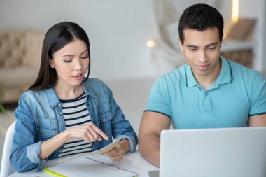 Koyu saçlı kadın ve esmer erkek masada oturuyor, kağıtlara bakıyor, dizüstü bilgisayardan bir şeyi kontrol ediyor.