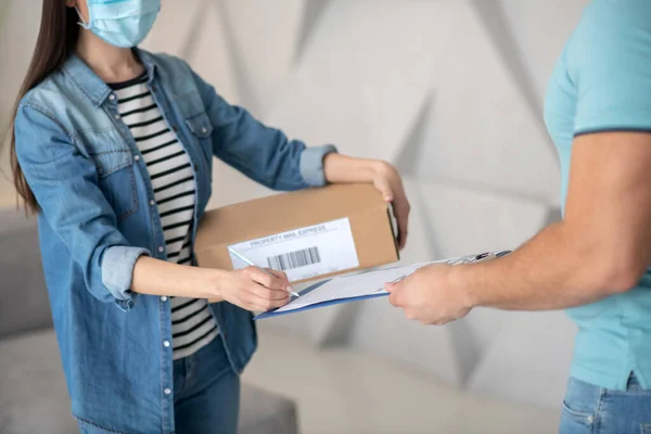 Mujer con una máscara protectora con una caja firmando un documento . — Foto de Stock