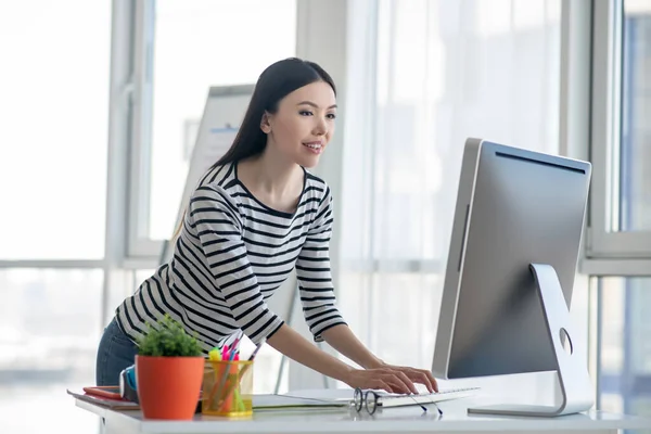 Donkerharige vrouw in een gestreept hemd bij de tafel en kijkend naar de computer — Stockfoto