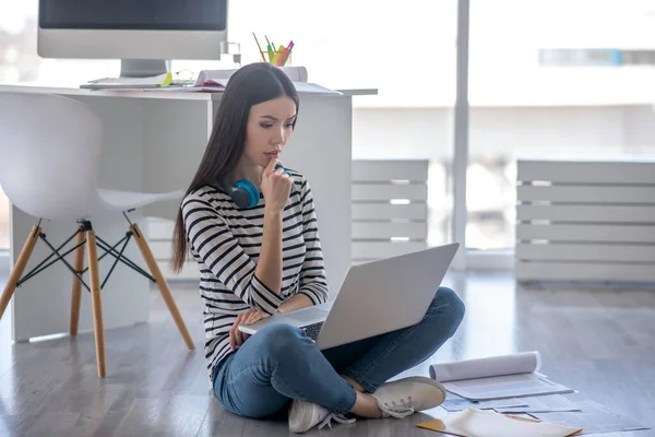 Menina de cabelos escuros com fones de ouvido trabalhando em um laptop e olhando pensativo — Fotografia de Stock