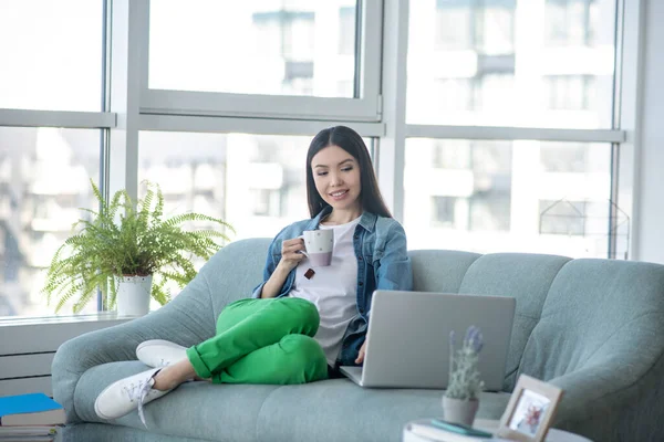 Giovane donna in giacca jeans seduta sul divano e guardando qualcosa online — Foto Stock