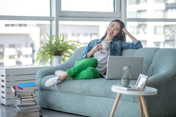 Giovane donna in giacca di jeans seduta sul divano e che prende il tè — Foto Stock