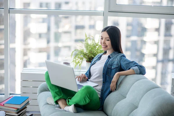 Ung kvinna i jeans jacka sitter på soffan och surfa internet — Stockfoto