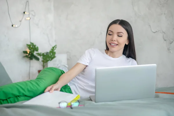 Joven mujer morena acostada en la cama y trabajando en el portátil — Foto de Stock