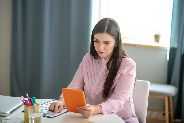 Mujer joven y bonita en una blusa rosa sentada a la mesa y sosteniendo una tableta — Foto de Stock