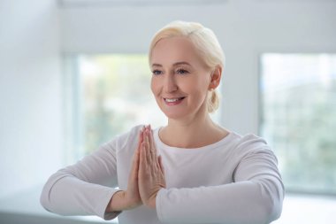 Olgun sarışın kadın namaste yapıyor, el ele tutuşuyor, gülümsüyor.